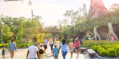 People walk along a sunny, tree-lined path at a zoo or theme park. Lush greenery, colorful flowers, and a large DreamWorks-inspired play structure resembling a treehouse create a lively, family-friendly atmosphere.