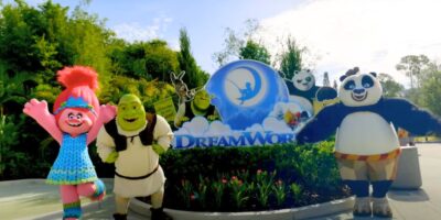 Costumed DreamWorks characters from Trolls, Shrek, and Kung Fu Panda pose and wave in front of a DreamWorks sign outdoors on a sunny day with trees in the background.