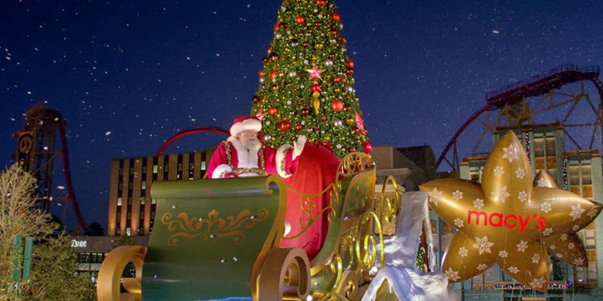Santa Claus sits in a green sleigh waving in front of a large, decorated Christmas tree and a glowing Macy's star, with universal holiday cheer spreading through the buildings and amusement park ride aglow in the night.