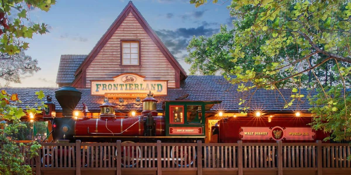 A red steam locomotive labeled "Walt Disney Railroad" is parked at the Frontierland Railroad Station, surrounded by wooden fences and green trees under a partly cloudy sky, capturing a classic Disney scene that feels far from vanishing.