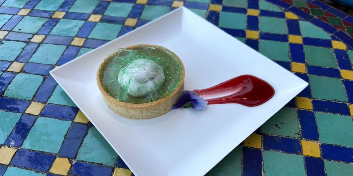 Green jelly tart with white topping and red sauce on a white plate, served at EPCOT International Festival's colorful tables.