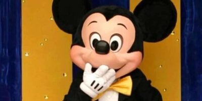 Mickey Mouse at Disneyland Resort playfully covers his mouth with a gloved hand, posing before a cheerful blue and yellow backdrop as numerous Disney attractions close down in 2026.