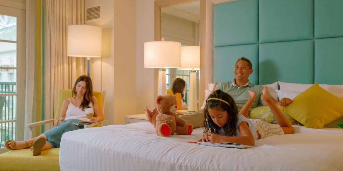 A family unwinds in their Disney Resort room; a child sketches on the bed as parents read after a magical day at the parks.