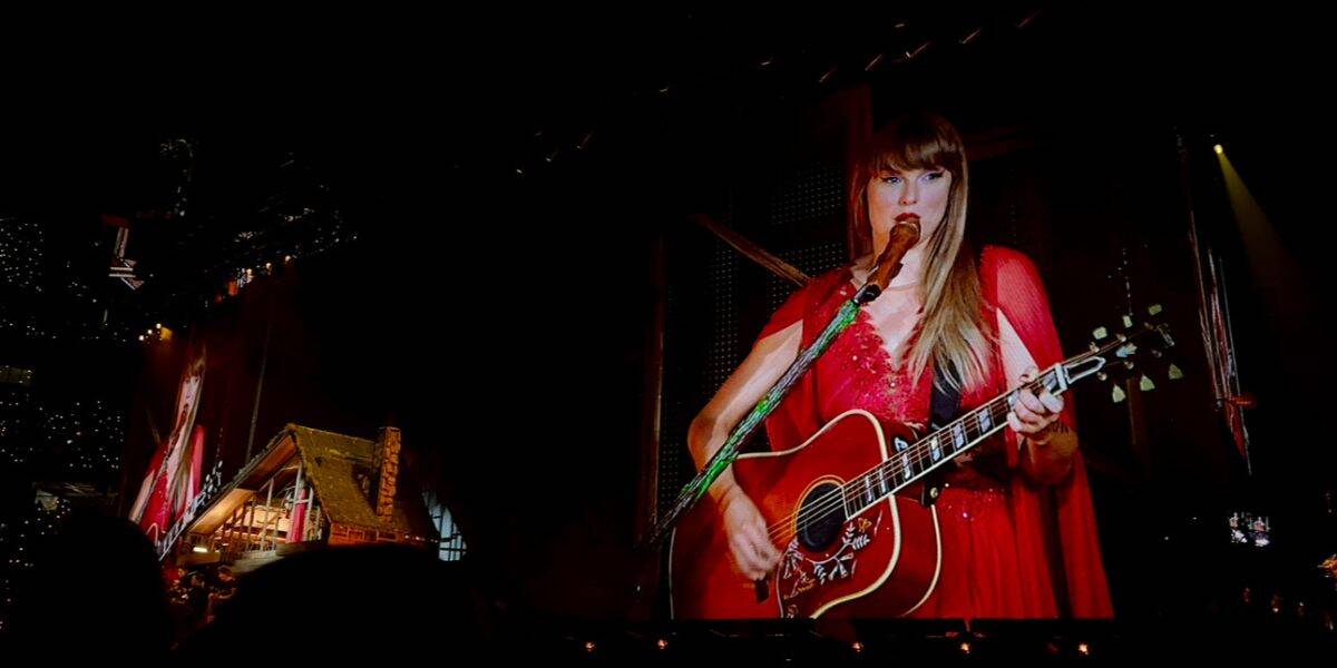 Taylor Swift dazzles onstage in a red dress, strumming guitar and singing, her performance magnified on giant concert screens.