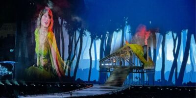 Taylor Swift in a green costume performs on stage before a forest set and big screen, evoking her Eras Tour magic for Disney+ fans.