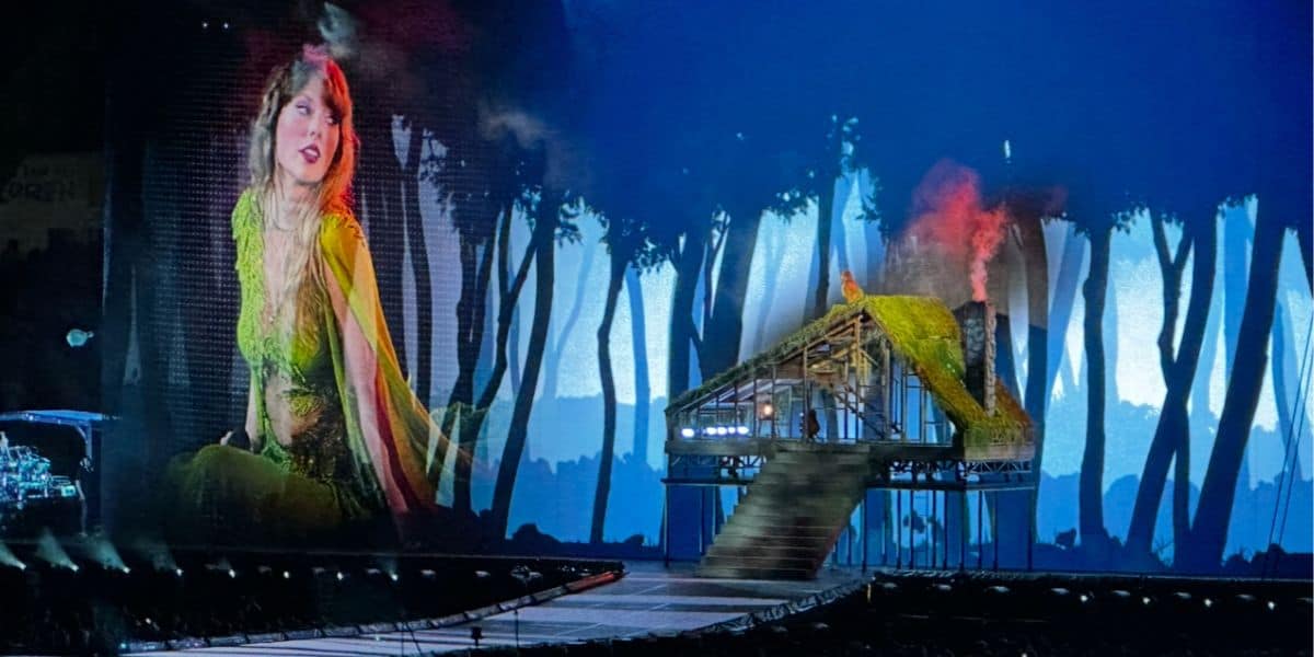 Taylor Swift in a green costume performs on stage before a forest set and big screen, evoking her Eras Tour magic for Disney+ fans.