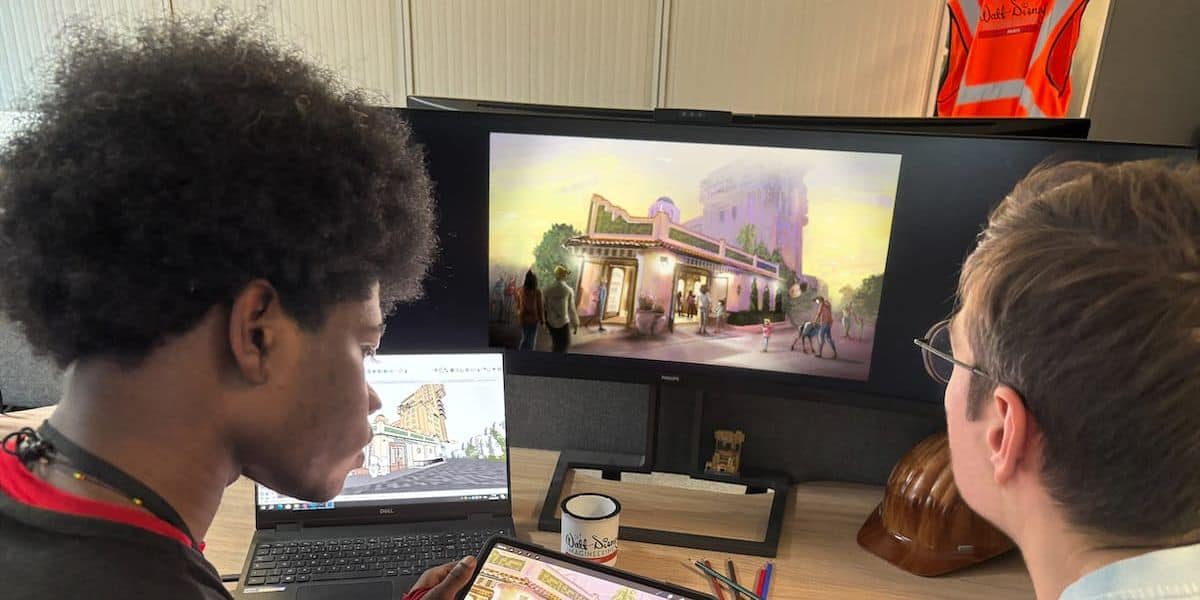 Two Imagineers review a theme park attraction blueprint on a computer monitor and tablet at their creative studio desk.