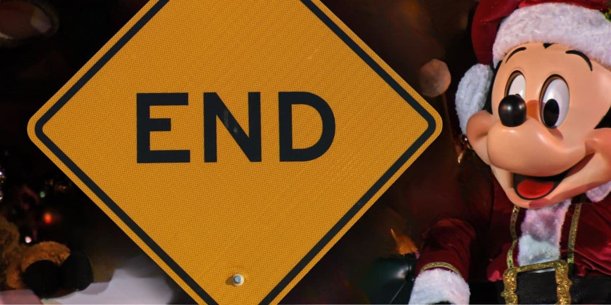 Mickey Mouse in a festive Santa suit stands by a yellow "END" road sign, ready to greet guests at Disney World.
