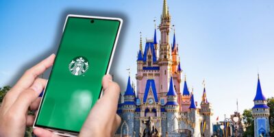 A guest using the Starbucks app on their phone with Cinderella Castle in the background under bright, sunny skies at Disney World inside Magic Kingdom for the 2026 Dining Plan.
