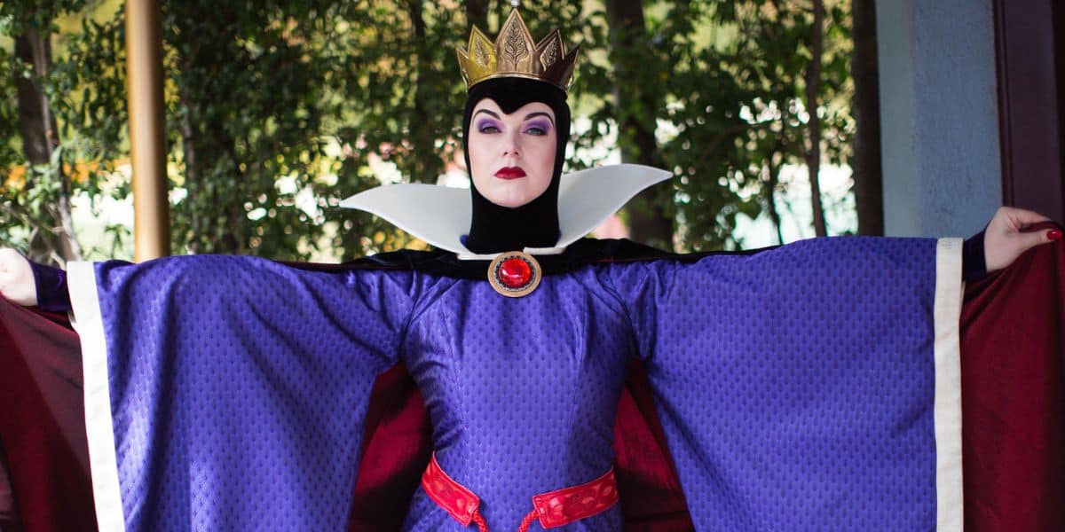 The Evil Queen from Snow White poses regally in her crown and purple robe, arms outstretched to greet park guests.