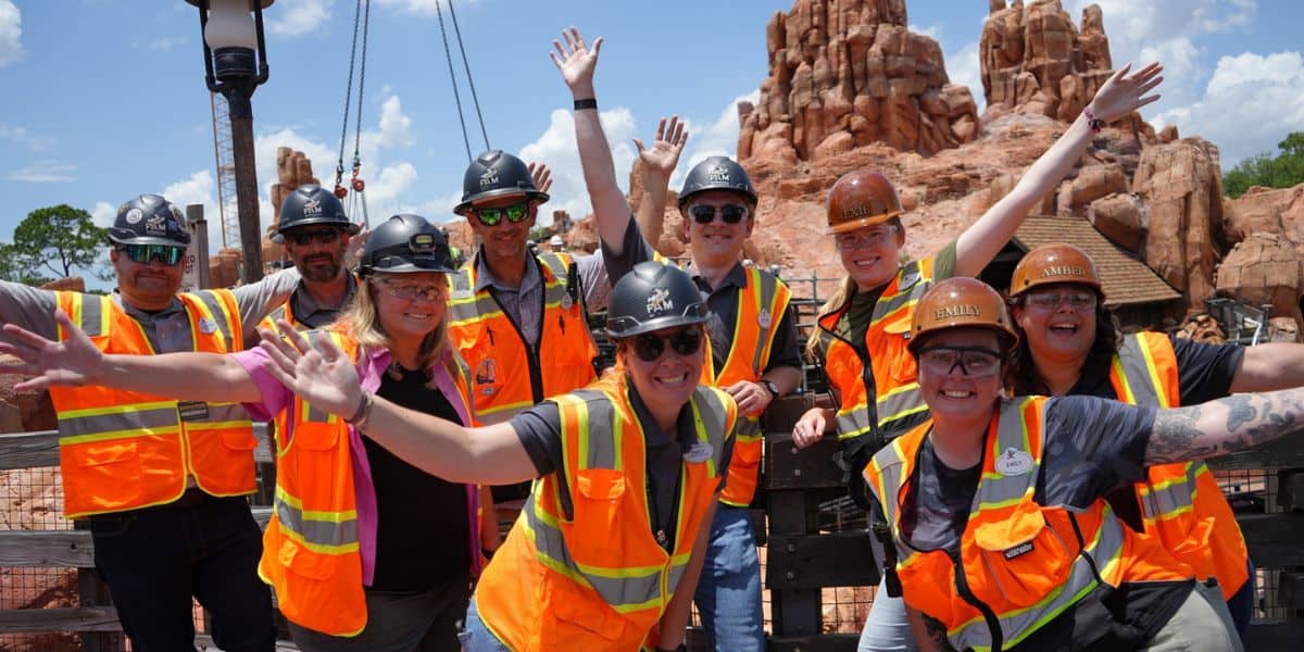 Construction crew in orange vests celebrates finished track, arms raised, ready for the Disney ride’s grand reopening.