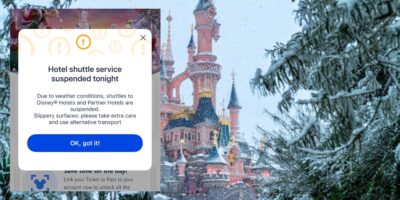 Sleeping Beauty Castle covered in snow with on-screen alert: hotel shuttle temporarily paused due to wintry weather conditions.
