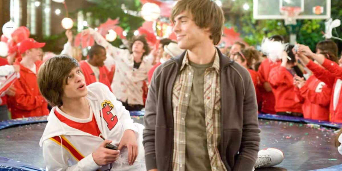Two teens chat on a trampoline during a spirited school carnival, as crowds in red cheer from behind like fans at peak festival hour.