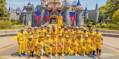 A baseball team in bright yellow uniforms poses at Magic Kingdom before Cinderella Castle, festooned with American flags.