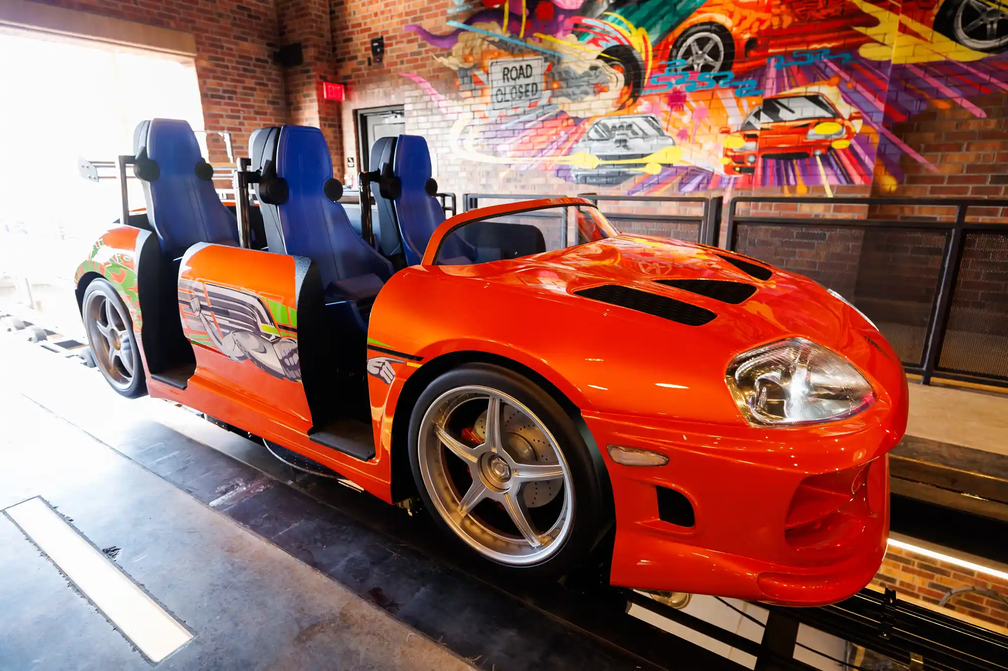 Sleek orange sports car ride vehicle with four seats, located indoors beside a vibrant mural in the arcade area.