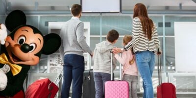 A family with suitcases checks an airport screen as Mickey Mouse welcomes them, hinting at an exciting Disney World adventure. Global Entry shutdown Disney travel
