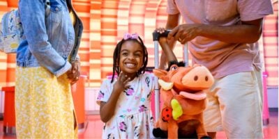 young guest with suitcase and Pumba stuffed animal waits in disney's hotel lobby with her parents