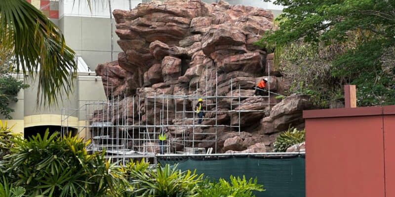 Crew members on scaffolding restore themed rockwork amid lush landscaping at an immersive attraction under construction which happens to be the Jurassic Park River Adventure at Universal Orlando.