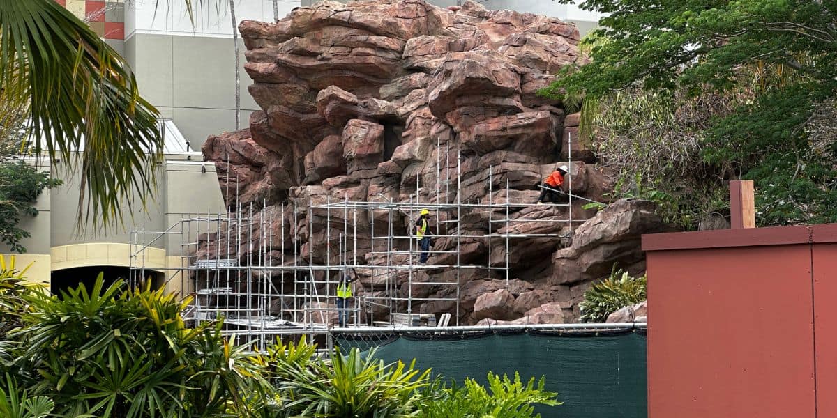 Crew members on scaffolding restore themed rockwork amid lush landscaping at an immersive attraction under construction which happens to be the Jurassic Park River Adventure at Universal Orlando.