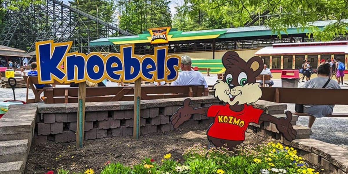 Knoebels entrance sign with mascot Kozmo welcoming guests, surrounded by benches, blooming flowers, and a coaster backdrop.