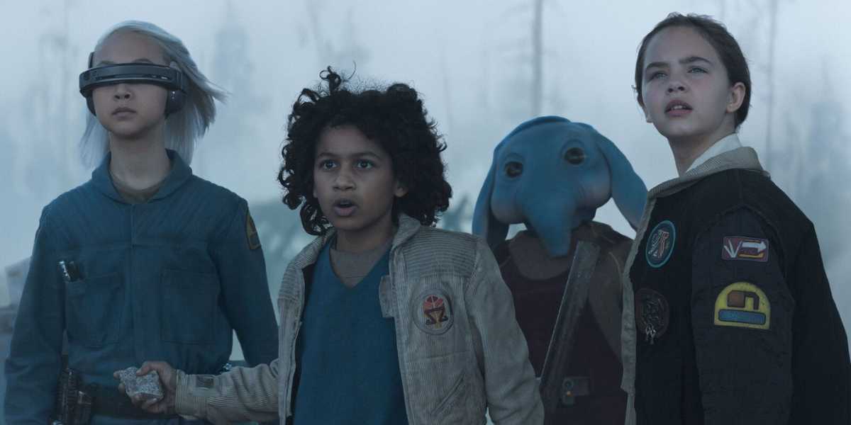 From left to right: KB (Kyriana Kratter), wearing a blue jumpsuit and a tech visor with short white hair, holds a small object in her hand; Wim (Ravi Cabot-Conyers), in a tan corduroy jacket with a patch emblem, stands wide-eyed at center; Neel (Robert Timothy Smith), a blue-skinned alien with large floppy ears, peers over the group from behind; and Fern (Ryan Kiera Armstrong), in a black bomber jacket adorned with colorful patches, gazes upward to the right