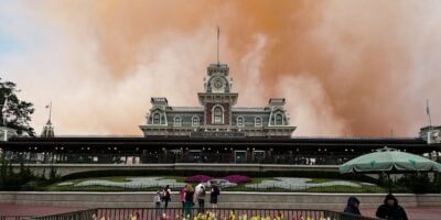 Guests gather outside the main entrance as vibrant orange smoke effects create a dramatic spectacle above the station façade at Magic Kingdom at Disney World. Saharan Dust Disney World