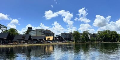 A serene lake mirrors lush trees and resort buildings under a sunny sky at Disney Orlando’s latest guest-exclusive retreat.