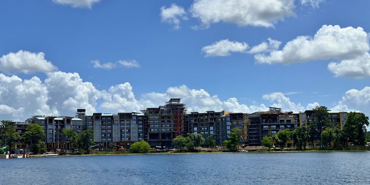A new Disney Orlando resort rises by a scenic lake, set to welcome park fans amid lush trees and blue skies.