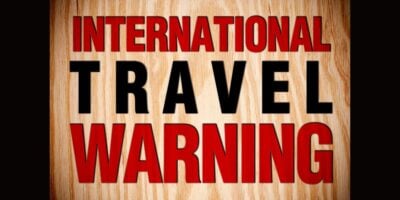 A wooden sign features bold red and black letters announcing "INTERNATIONAL TRAVEL WARNING," resembling the style of themed park advisories often seen at attractions or entrance gates as new Disney travel warnings are sent out by the State Department.