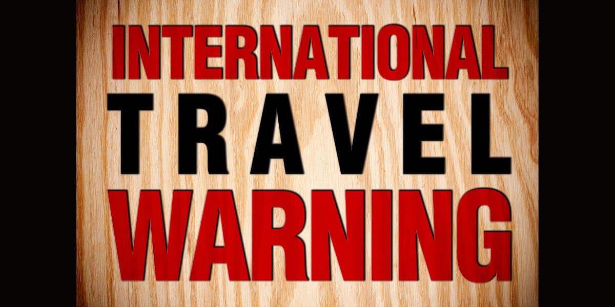 A wooden sign features bold red and black letters announcing "INTERNATIONAL TRAVEL WARNING," resembling the style of themed park advisories often seen at attractions or entrance gates as new Disney travel warnings are sent out by the State Department.