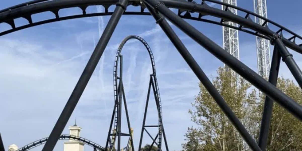 Soaring black steel coaster track twists through thrilling loops and turns, framed by blue skies and lush trees—get ready for heart-pounding excitement at the amusement park!.