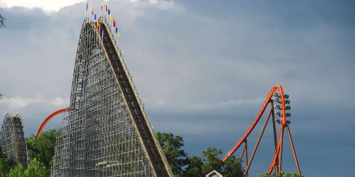 Classic wooden and towering steel roller coasters, each crowned with fluttering flags, soar above lush green trees beneath a dramatic cloudy sky—capturing the excitement of Six Flags-style thrills and the cutting-edge operations that define top American amusement parks.