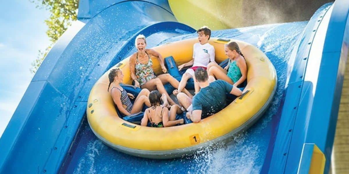 Six guests speed down the iconic blue Rapids Racer slide at Six Flags, laughing together on a bright yellow raft as they experience one of the park’s most thrilling water attractions.