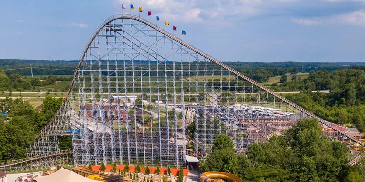 The iconic wooden roller coaster at Six Flags soars above lush treetops and the bustling parking lot, topped with vibrant flags, capturing the nostalgic excitement and flawless operations that make American theme parks unforgettable.