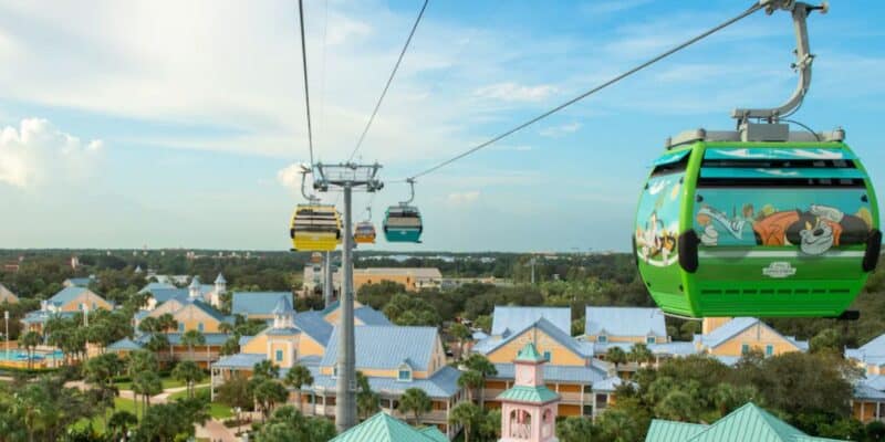 Disney Skyliner gondolas glide high above the vibrant Disney World Moderate Resort, offering guests panoramic views of blue-roofed resort buildings and lush palm trees beneath a bright, sunny sky.