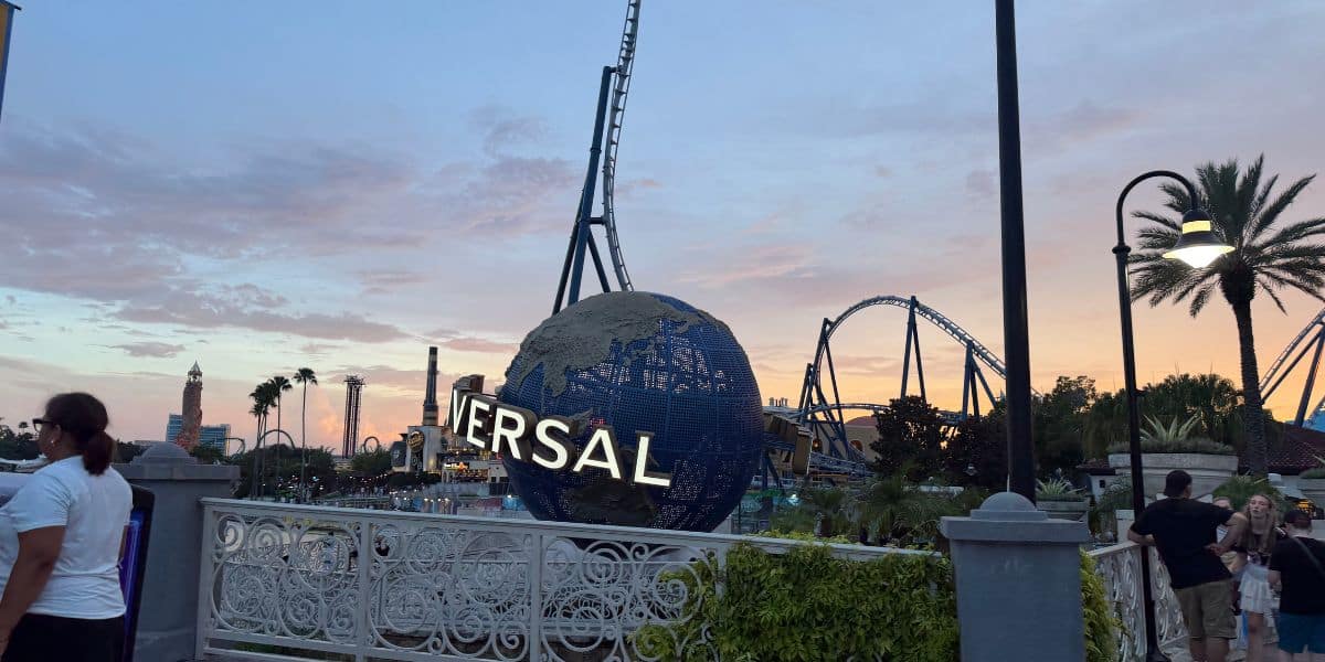 The iconic Universal globe welcomes guests as roller coasters soar in the golden sunset, with visitors strolling along lively pathways lined with palm trees.