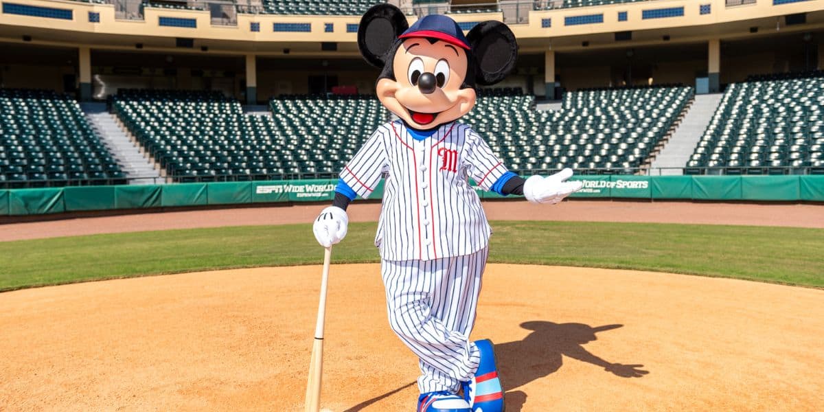 Mickey Mouse dons his classic baseball uniform and poses with a bat on a lively Disney ballfield, with rows of empty stadium seats creating an exciting anticipation for fans.
