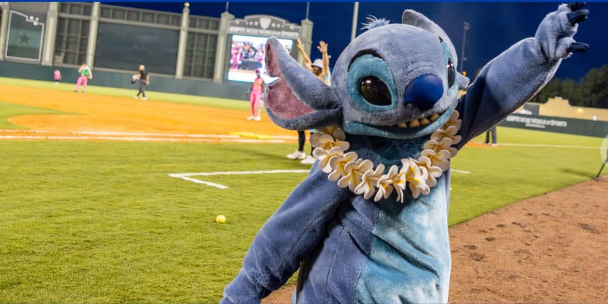 Stitch wearing a colorful lei greets fans with a wave on the baseball field, as the scoreboard and athletes create an exciting game-day atmosphere in the background.