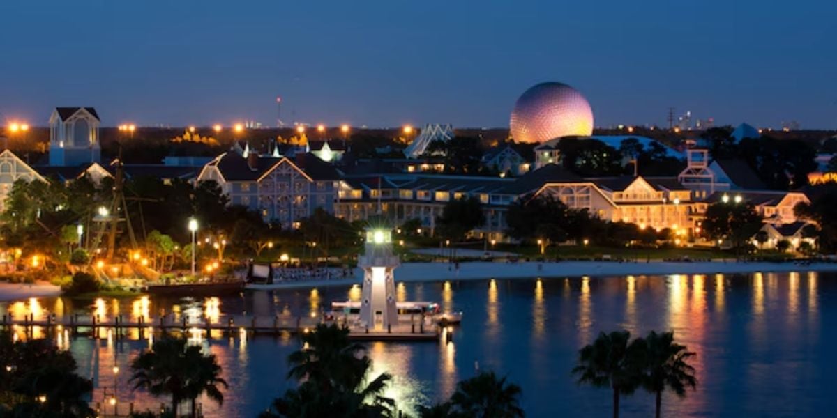 Nighttime scene of a lively resort area reminiscent of a theme park, featuring an illuminated dome and entertainment pavilions glowing along the waterfront, their colorful lights shimmering in the water below.