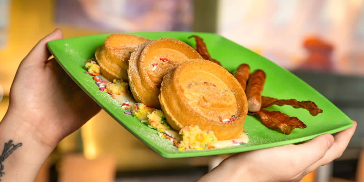 A stack of funfetti pancakes, crispy bacon, and sausage links served on a green plate held by eager hands, just like a breakfast treat you’d enjoy at Universal Studios’ family dining spots.