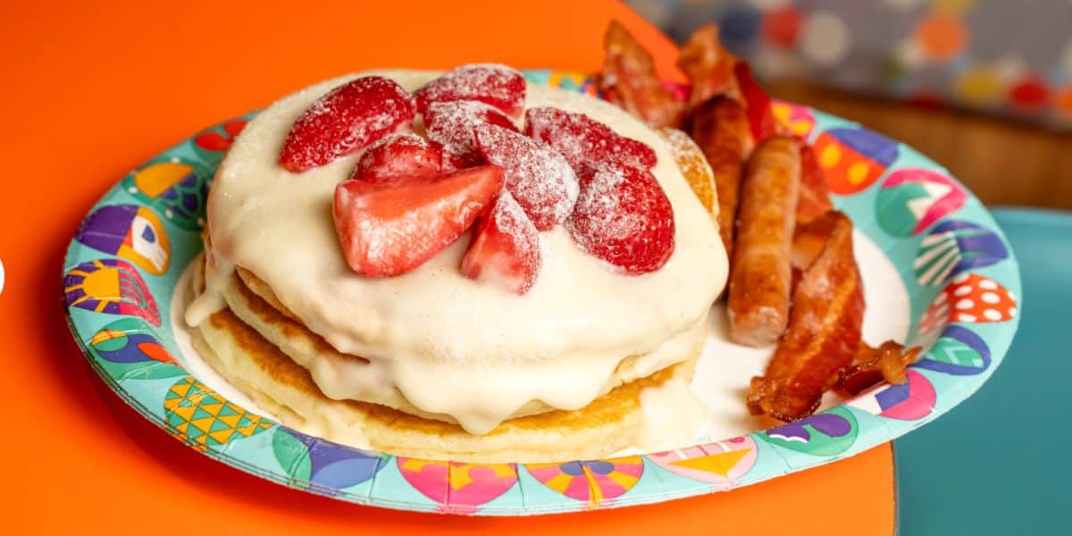 A stack of three fluffy pancakes crowned with cream and strawberries, alongside crispy bacon and savory sausage, all served on a vibrant plate bursting with color—perfect for fueling up before a day of adventure at your favorite theme park.