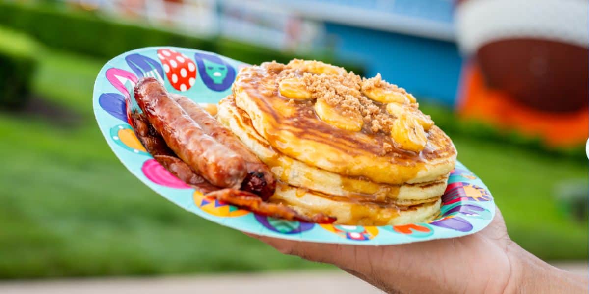A guest enjoys classic buttermilk pancakes topped with banana slices and syrup, served with two sausage links on a plate at Main Street’s charming breakfast café in Magic Kingdom.