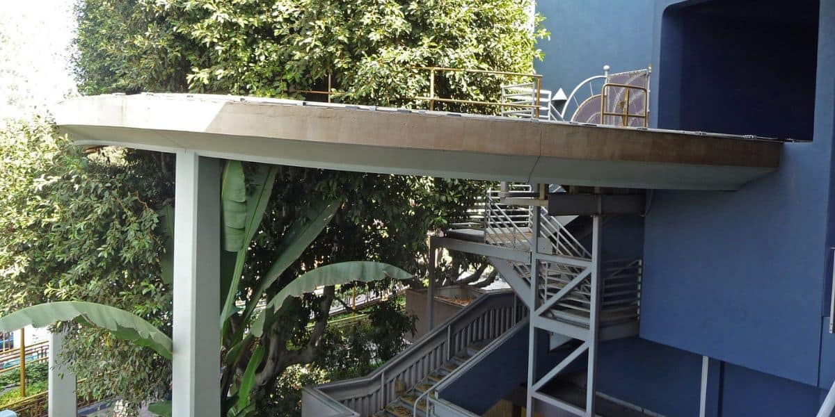 A thrilling elevated walkway seems to vanish into thin air, dramatically held up by a single pillar, with winding stairs below and vibrant greenery enhancing the adventurous theme park atmosphere.