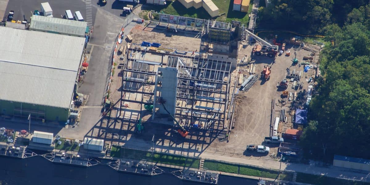Aerial view of a bustling construction zone featuring steel framework, cranes, and busy crew members set among greenery and nearby buildings, unveiling what could be the beginnings of an exciting new Disney Parks attraction or mysterious structure taking shape for future guests.