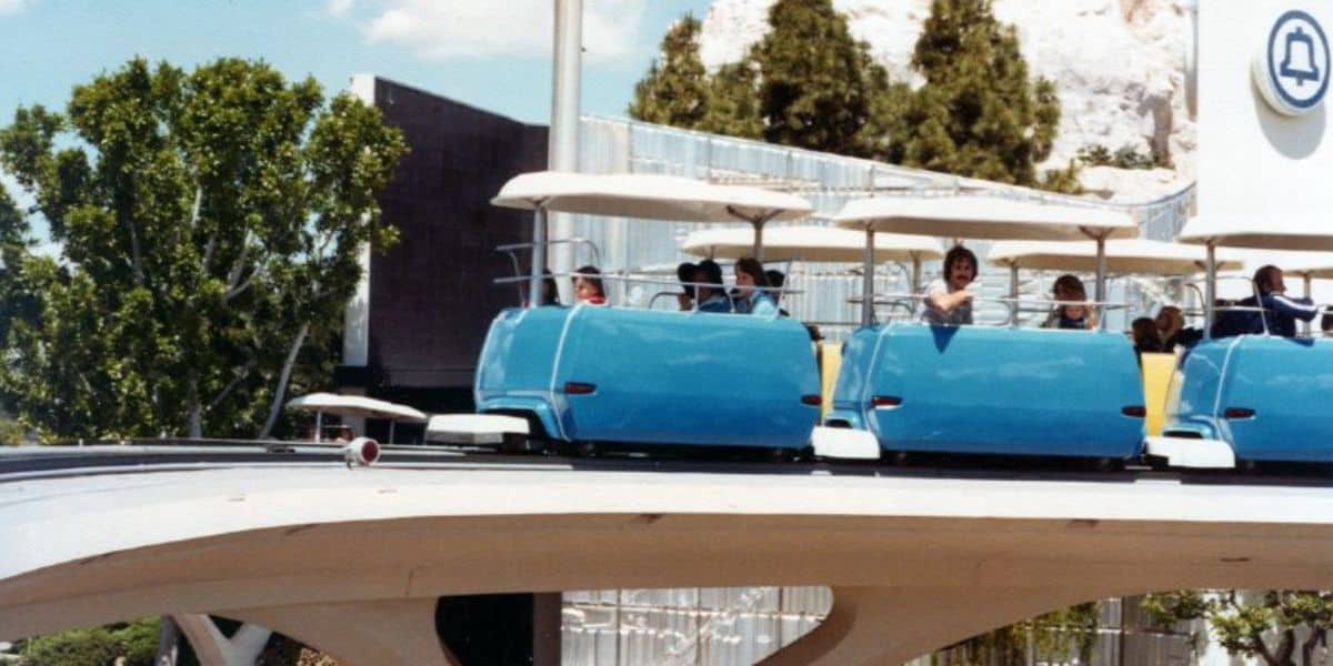 Guests zoom along Tomorrowland Speedway in signature blue cars on an elevated track at Magic Kingdom, surrounded by lush trees and iconic park architecture.