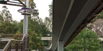 Beneath the sleek beams of a theme park monorail, lush trees line the walkway as guests ascend wide stairs with sturdy railings. A glowing ring light mounted on a nearby pole adds extra flair to this lively entertainment hub.