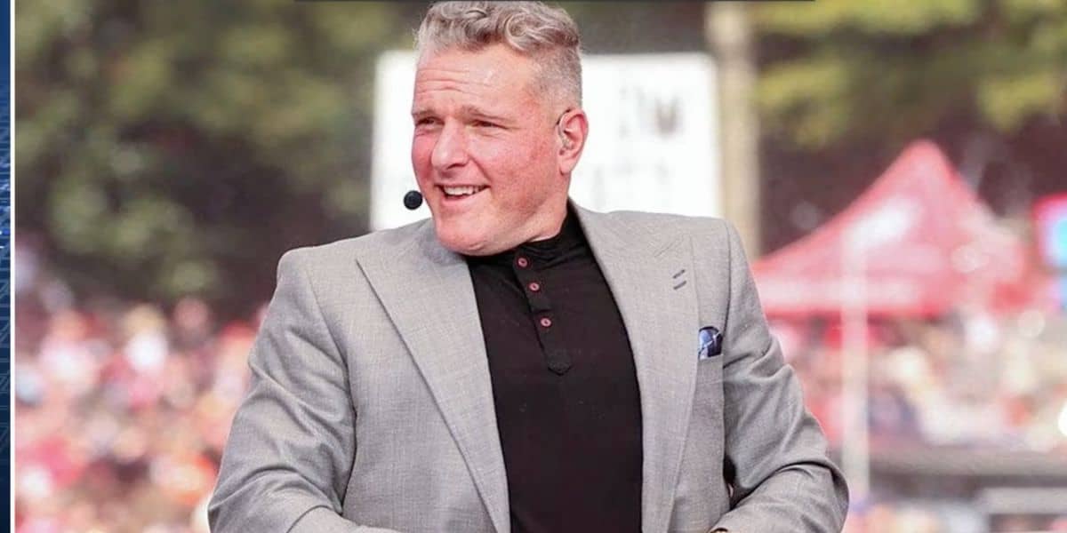 Pat McAfee, dressed in a gray suit jacket and wearing a headset, beams with excitement while sitting outdoors among the buzzing event crowds—capturing the lively energy of a theme park broadcast as fans anticipate his take on the latest Disney drama.