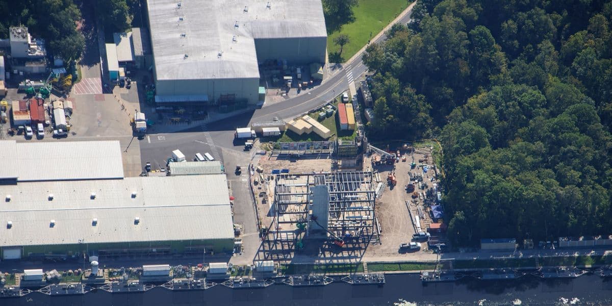 Aerial view of a mysterious new attraction under construction, featuring an intricate steel framework and surrounded by park buildings, lush landscaping, and a winding guest roadway.