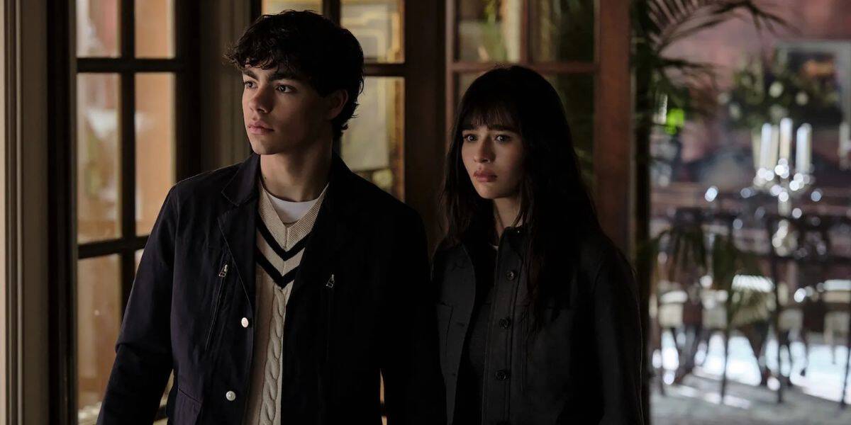 Two teens stand inside an immersive theme park attraction, their expressions serious as sunlight filters through themed set windows behind them.