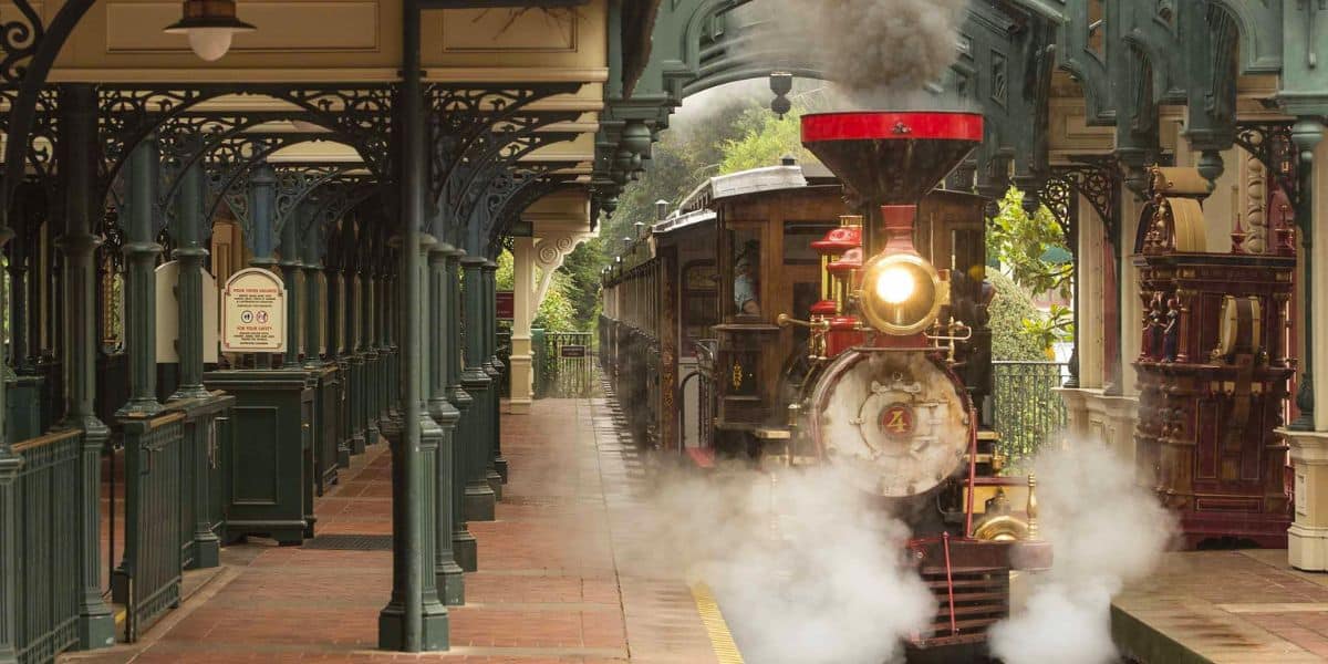 A vintage steam train pulls into Main Street Station at Disneyland, billowing white steam as excited guests look on from the platform.